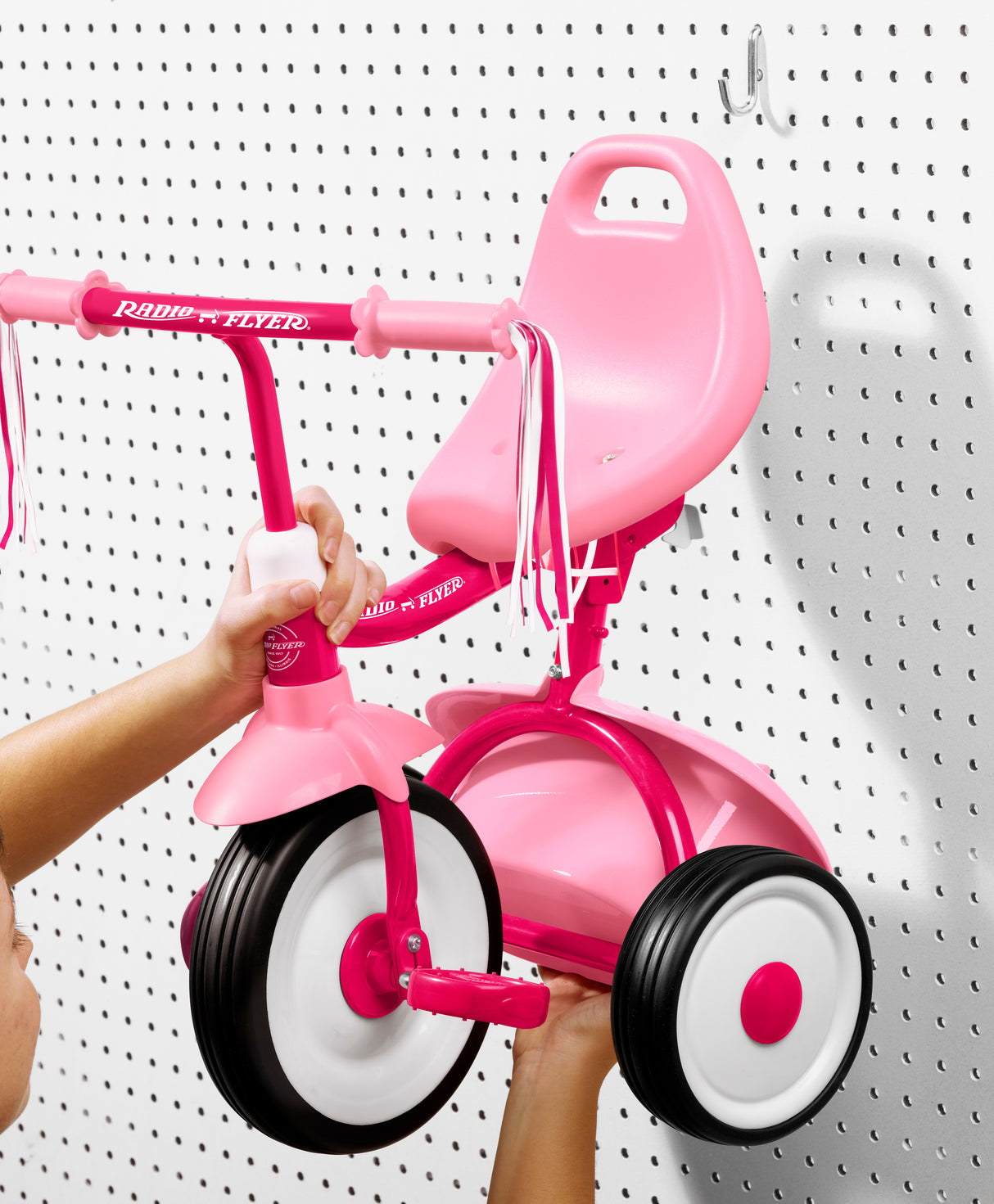 Pink folded Radio Flyer tricycle held by a person hanging it up on a white pegboard background