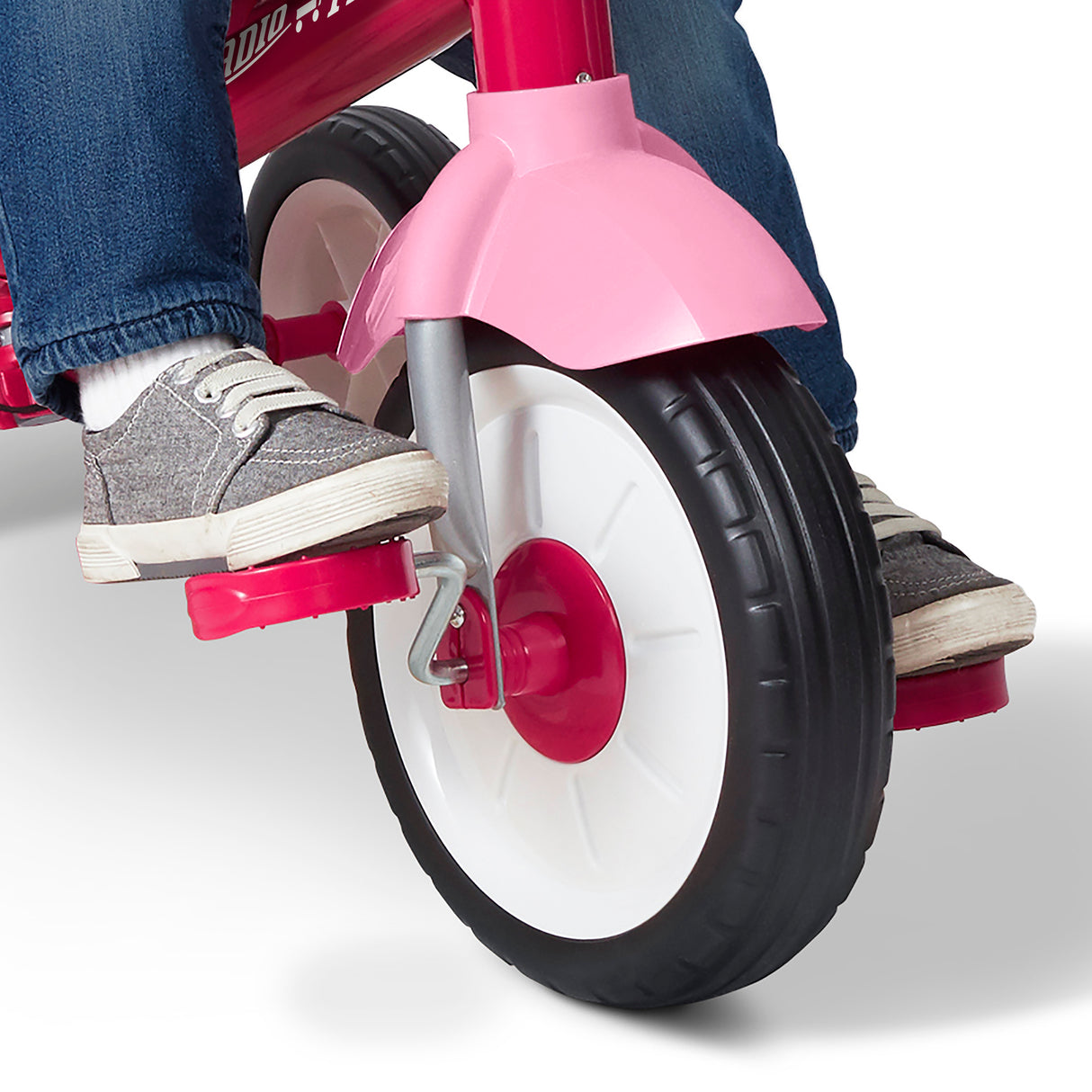 Close-up of a child riding a pink tricycle, showing the front wheel, pink fender, foot pedals, and the rider's feet in gray canvas sneakers and blue jeans against a white studio background.
