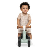 A smiling toddler with curly hair wearing beige and white overall shorts sits on a mint green balance bike with brown handlebars against a solid white background.