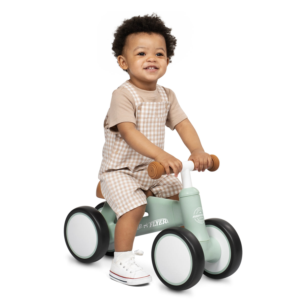 A smiling toddler wearing a beige and white overall shorts sitting on a mint green wooden balance bike with white wheels and brown handle grips, photographed against a clean white background.