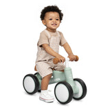 A smiling toddler wearing a beige and white overall shorts sitting on a mint green wooden balance bike with white wheels and brown handle grips, photographed against a clean white background.