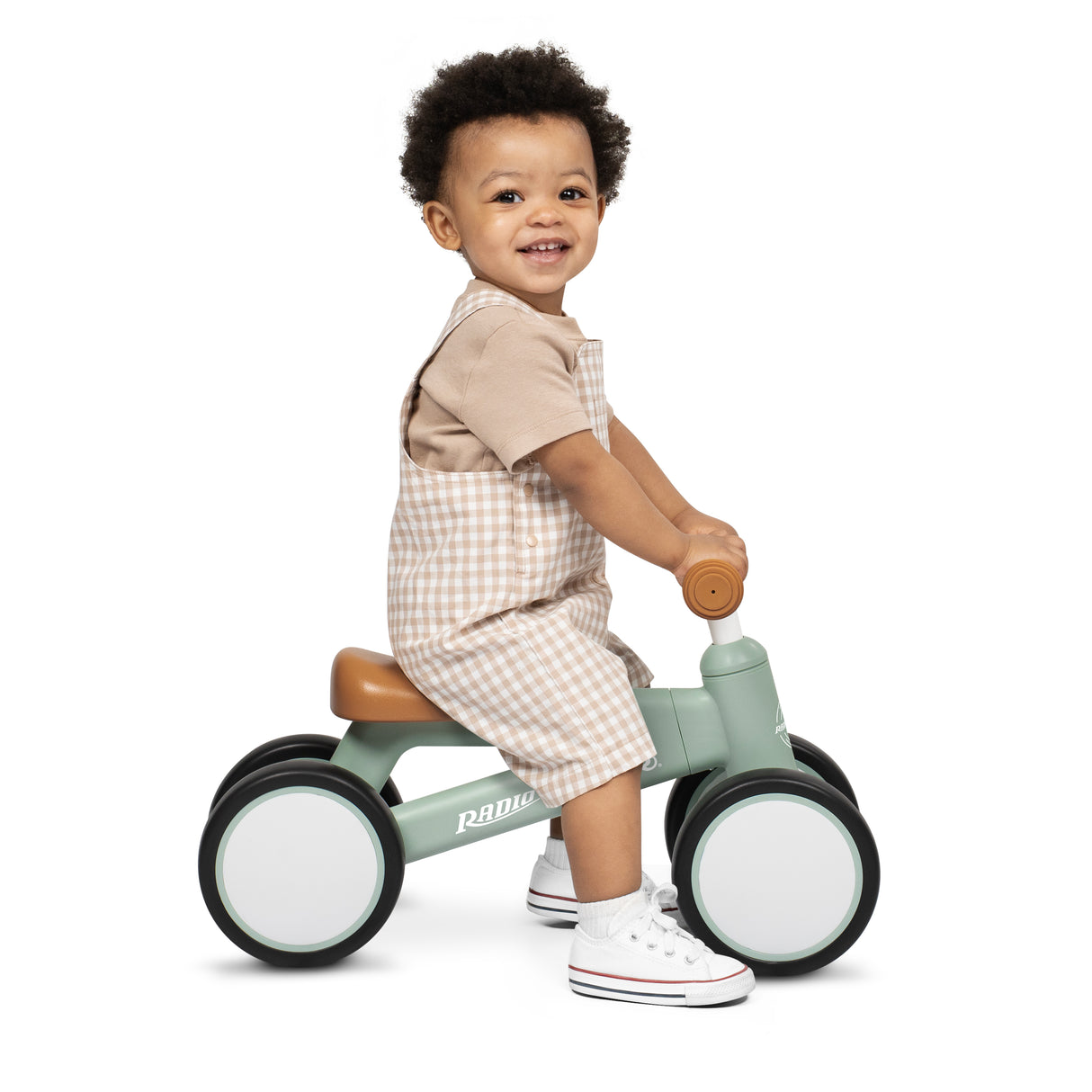 A toddler sitting on a Radio Flyer balance bike, wearing beige and white overall shorts and white sneakers, photographed against a solid white background.
