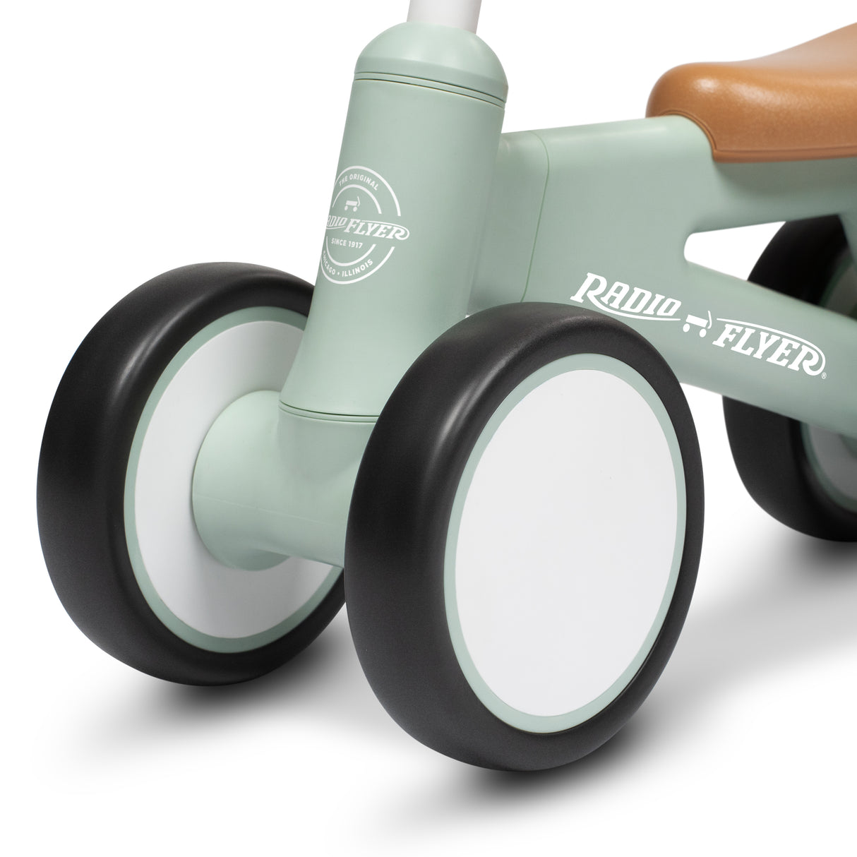 Close-up of a Radio Flyer balance bike showing the mint green frame, black and white wheels, and a brown saddle on a white background.