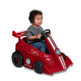 Young child smiling while riding the red Grow With Me Racer with hands on the steering wheel and seat belt fastened