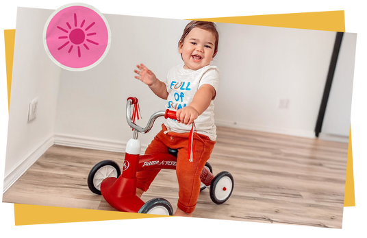 Child riding a red tricycle indoors with a pink sun sticker overlayed. 